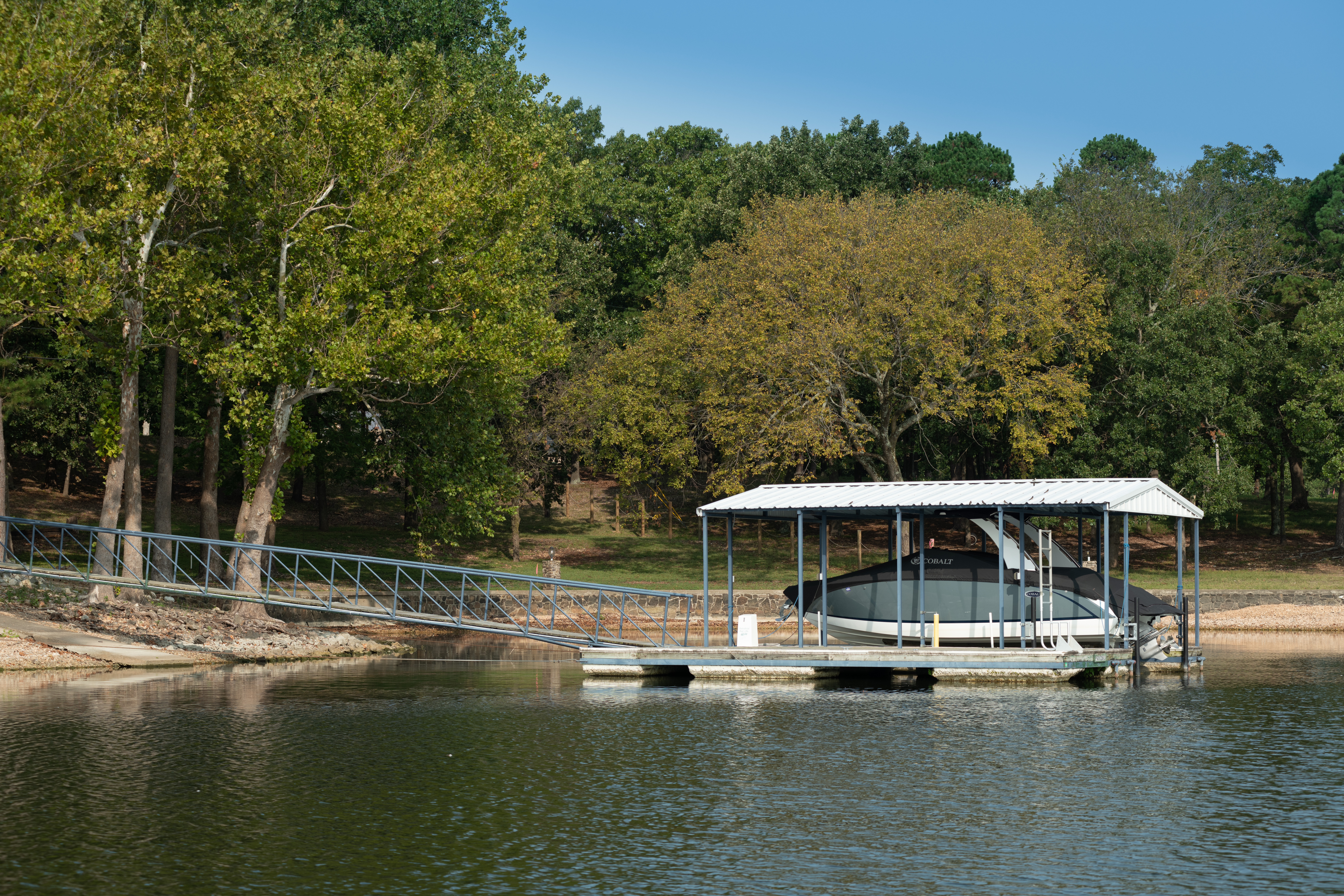 Covered boat dock with a boat lift and walkway on a tree-lined lake.