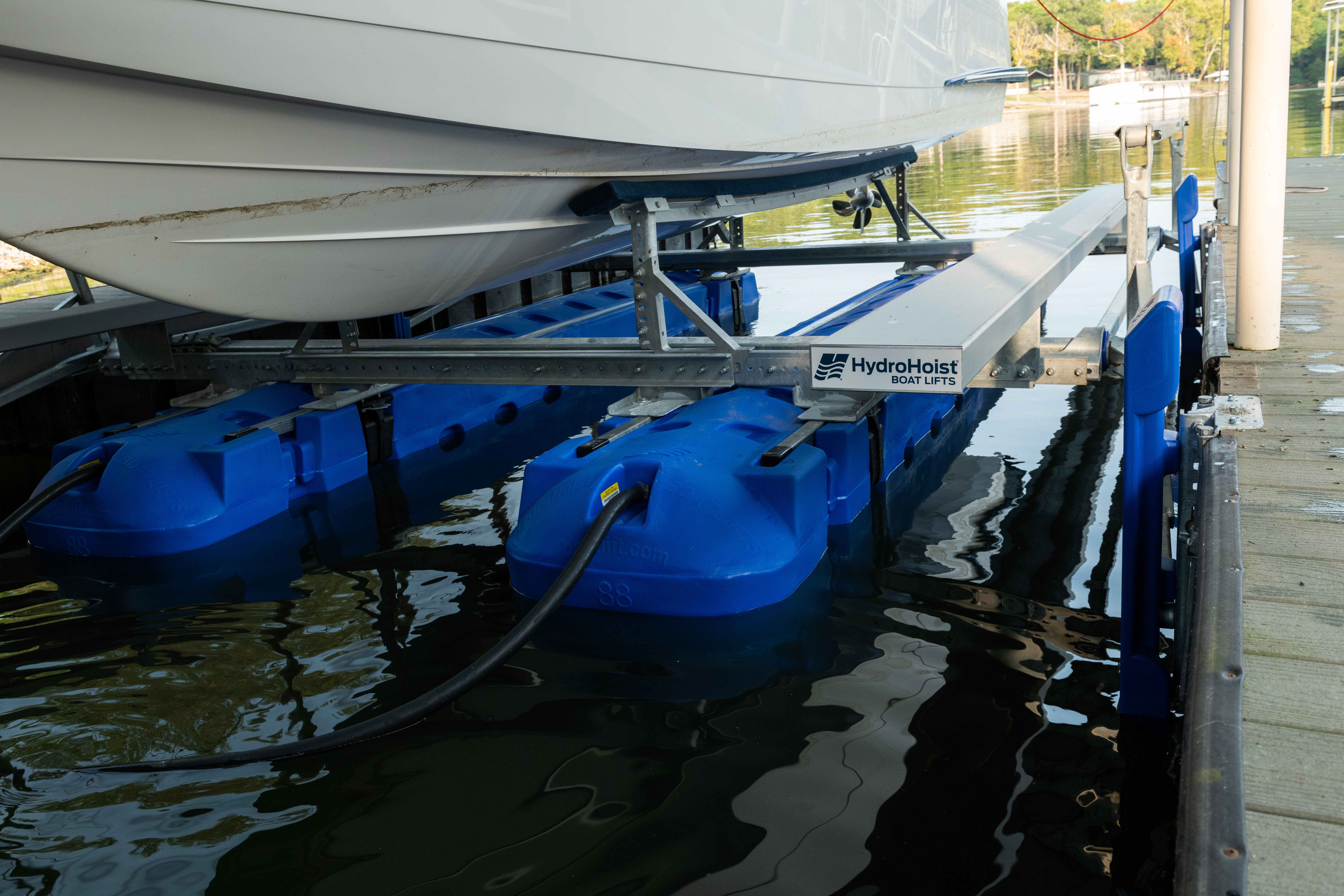 A white boat is secured on a blue HydroHoist boat lift in the water, with the logo visible on a metal frame.